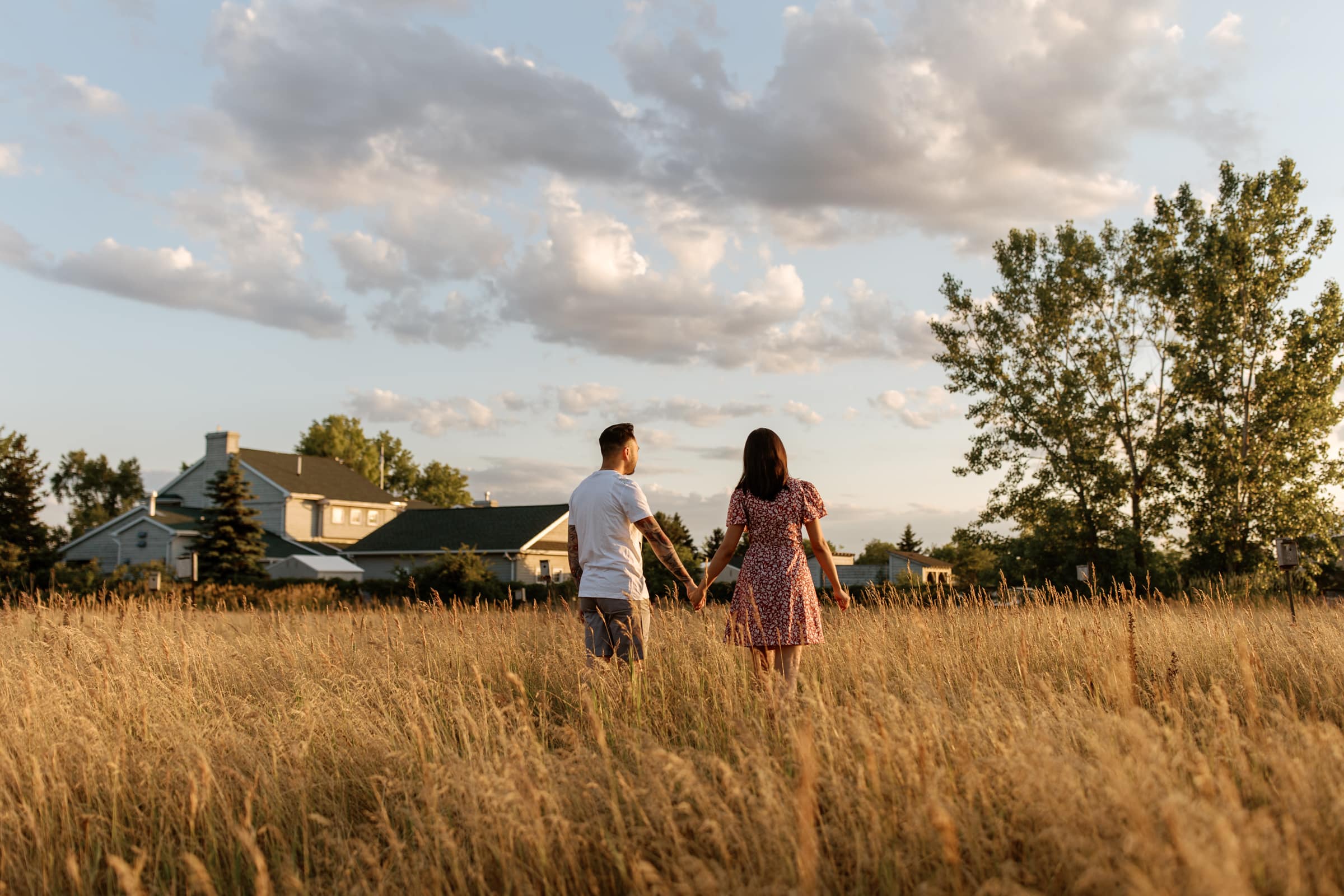 A couple enjoying a leisurely stroll through a picturesque field with a charming house in the background during their golden hour engagement session in Toronto.