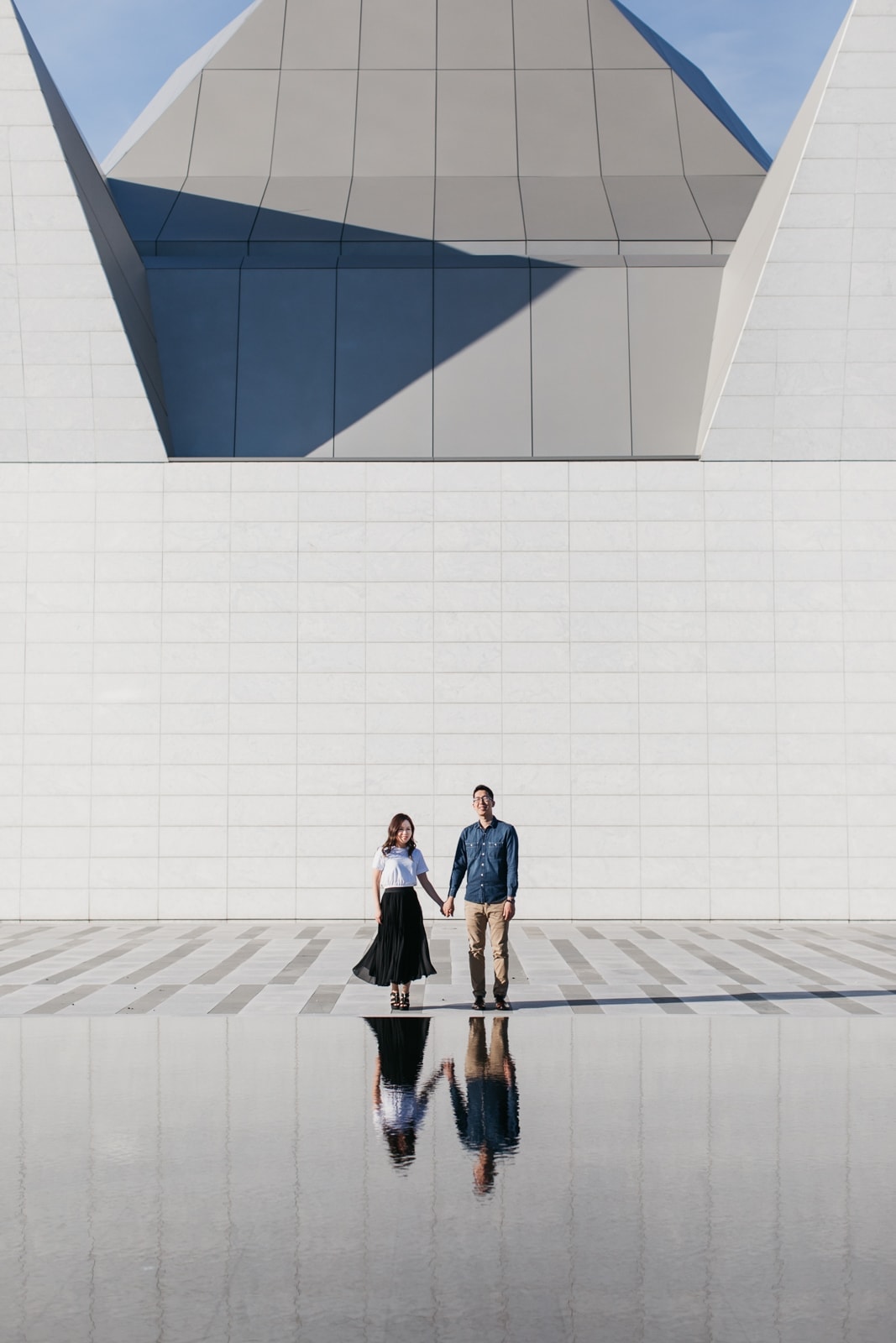 A couple stand holding hands in front of the Aga Khan Museum with a reflection in a mirrored pool during their engagement session.