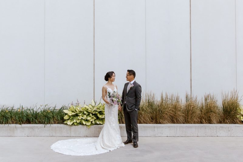 A bride and groom stand hand in hand while looking at each other in front of a clean white wall and flower bed with tall grass.