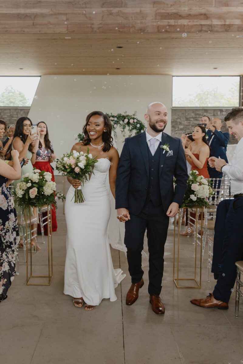 A bride and groom walk down the aisle during their wedding recessional, while guests blow bubbles in the Gazebo at their Guild Inn Estate wedding in Toronto