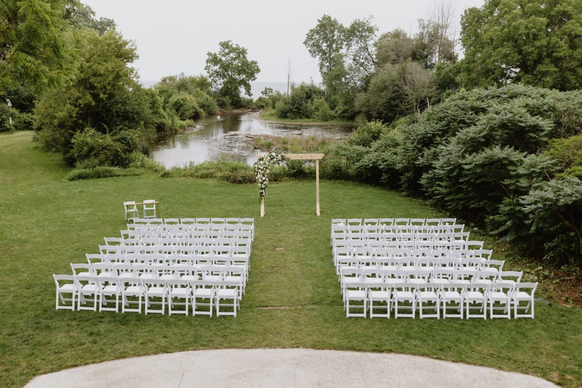 A beautiful outdoor wedding ceremony set up on Harding Waterfront Estate's grounds, featuring a white floral arch, elegant white chairs overlooking lake Ontario and and bordered by Joshua’s Creek.