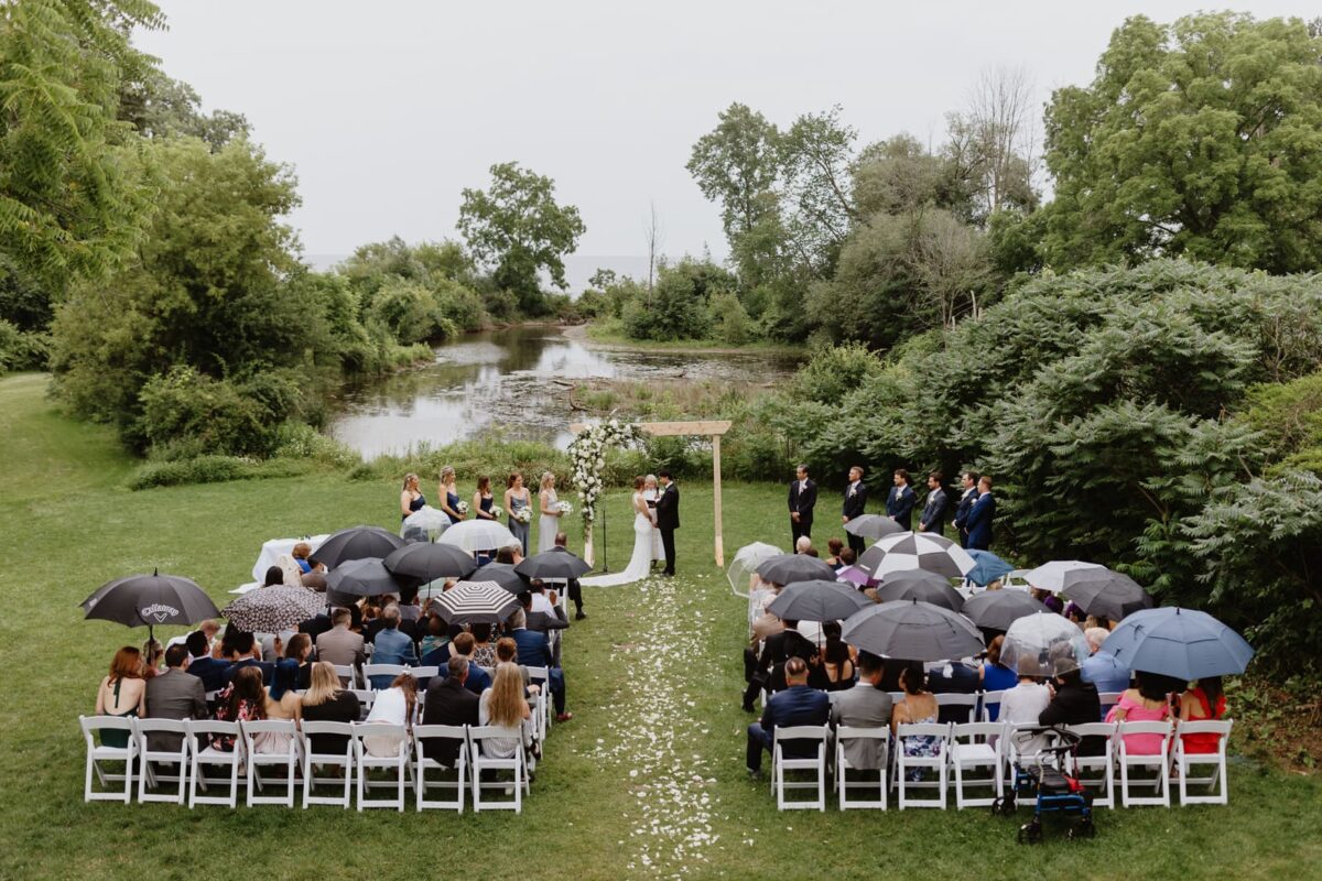 Bride and Groom exchange vows during outdoor wedding ceremony at Harding Waterfront Estate on a rainy day, overlooking lake Ontario and and bordered by Joshua’s Creek.