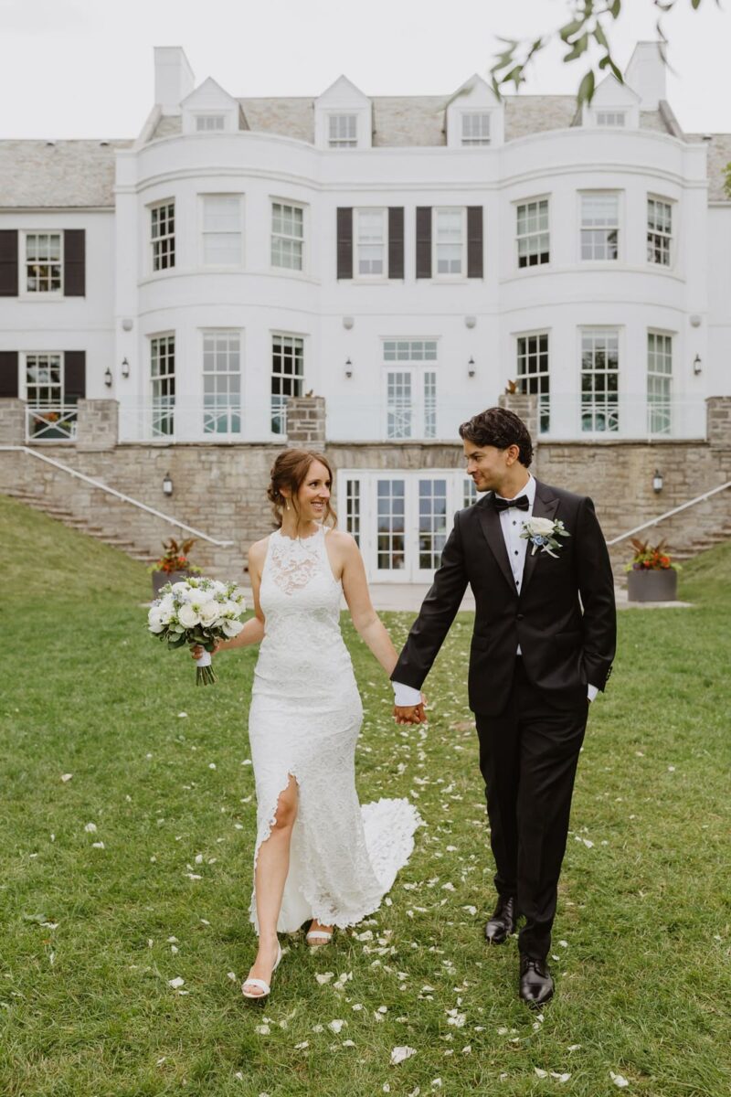 A bride and groom walking in front of the Bell-Gairdner House at Harding Waterfront Estate in Mississauga, a beautiful white mansion where their wedding took place.