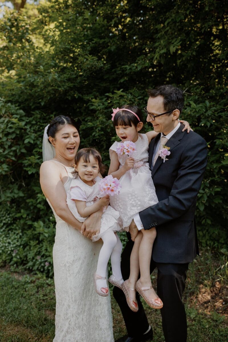 A family captured in wedding photos, with the bride and groom holding their two young daughters during a playful candid moment.