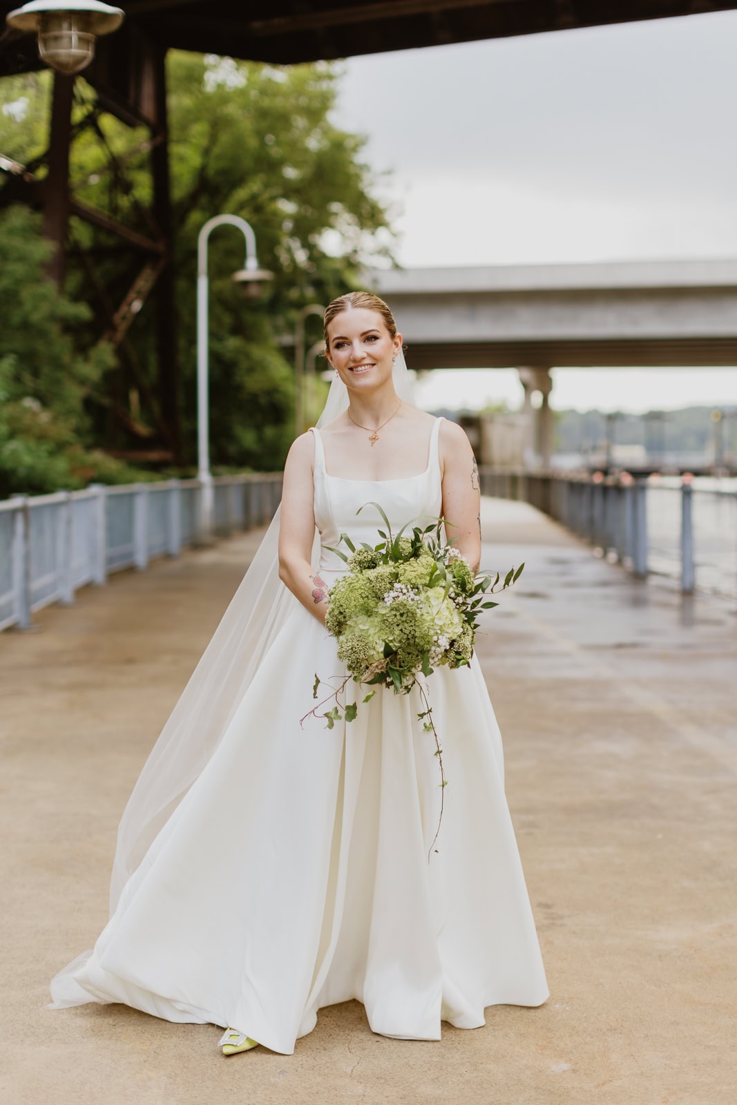 A bride in a white Jenny Yoo wedding dress smiling, holding a large green bouquet, on a path under a bridge.