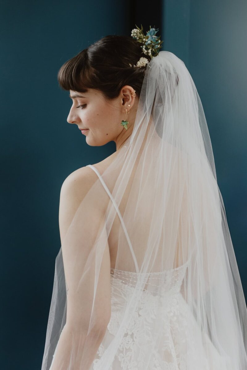 A bride wears a Stella York wedding dress with a delicate veil adorned with small flowers, with a subtle smile, captured in a profile view against a dark blue background.