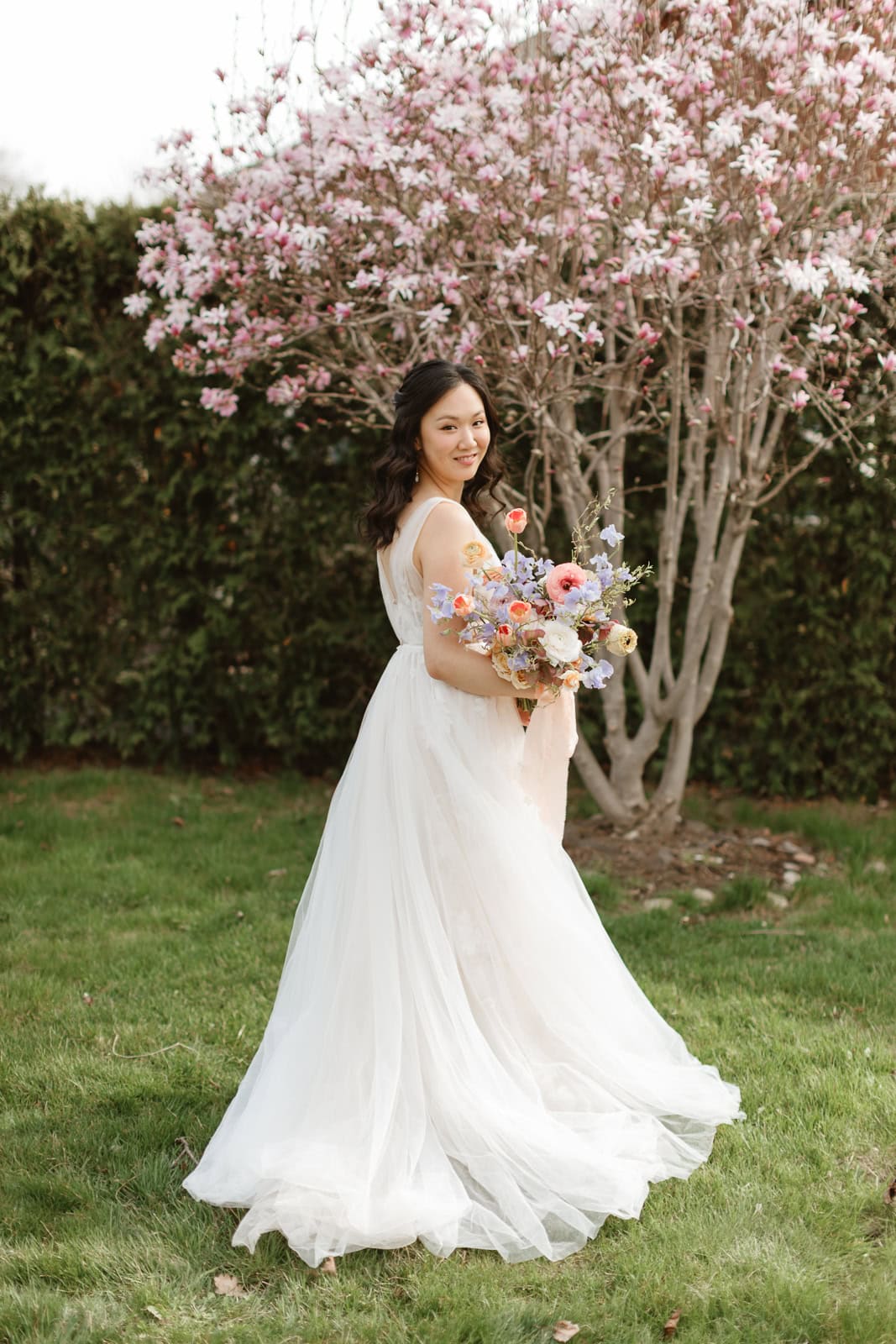 A bride in a flowing white Willoby by Watters wedding gown holds a colorful spring bouquet, smiling in a garden with a blooming pink magnolia tree.